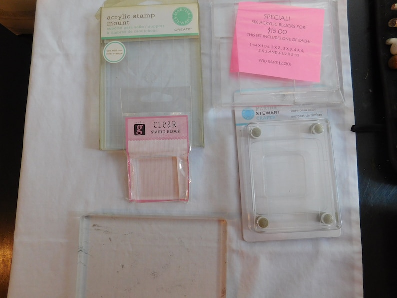 Puede incluir: Un conjunto de seis bloques de sellos de acr&iacute;lico transparente de diferentes tama&ntilde;os, empaquetados en una bolsa de pl&aacute;stico transparente con una etiqueta rosa que dice "SPECIAL! SIX ACRYLIC BLOCKS FOR $15.00 THIS SET INCLUDES ONE OF EACH 1X1, 1X2, 2X2, 3X4, 4X4, 5X2 AND 4X2, 5X2, 5X5 YOU SAVE $2.00". Tambi&eacute;n hay dos soportes de sellos de acr&iacute;lico transparente, uno con una etiqueta rosa que dice "CLEAR stamp stock" y el otro con una etiqueta blanca que dice "acrylic stamp mount".