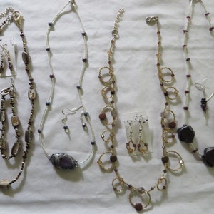 May include: Five sets of necklaces and earrings. The necklaces are made with various materials, including silver, gold, and brown beads. The earrings are made with silver, gold, and brown beads.