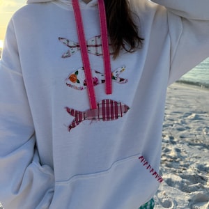 May include: White hoodie featuring three fish appliqués made from patterned fabrics. The fish are in pink, red, and floral patterns. The hoodie has a drawstring with pink ribbon and a kangaroo pocket. The pocket stitching is pink.