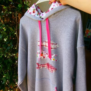 May include: Gray hooded sweatshirt with a floral-patterned hood lining and pink drawstrings. The sweatshirt features three decorative fabric fish appliqués in floral and plaid patterns. A pink-stitched pocket is visible on the front.