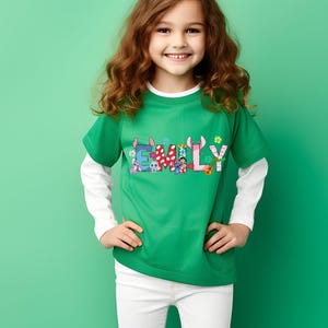 May include: A green t-shirt with the name "EMILY" in colorful letters, featuring cartoon characters and floral accents. The shirt has short sleeves and a white long-sleeved undershirt. The person is wearing white pants and is smiling.