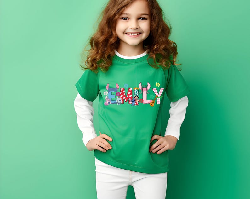May include: A green t-shirt with the name "EMILY" in colorful letters, featuring cartoon characters and floral accents. The shirt has short sleeves and a white long-sleeved undershirt. The person is wearing white pants and is smiling.