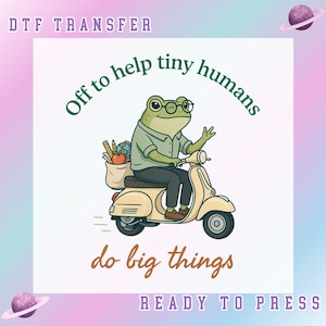 May include: A DTF transfer featuring a green frog wearing glasses, riding a beige scooter. The frog carries a bag with a ruler, pencil, and globe. Text reads: DTF TRANSFER, Off to help tiny humans, do big things, READY TO PRESS. Includes planet icons.