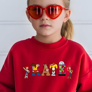 May include: A red sweatshirt with the word "SKATE" in colorful cartoon letters, each featuring a character from a popular animated movie. The person in the image is wearing heart-shaped red sunglasses.