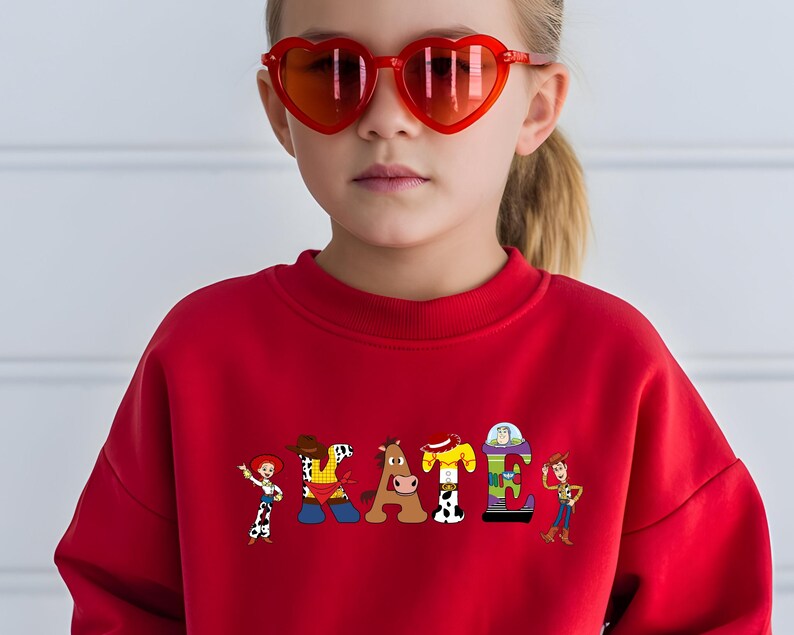 May include: A red sweatshirt with the word "SKATE" in colorful cartoon letters, each featuring a character from a popular animated movie. The person in the image is wearing heart-shaped red sunglasses.
