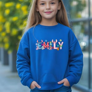May include: A royal blue sweatshirt with the name "EMILY" in colorful, whimsical letters. The letters are decorated with various cartoon characters and floral accents. The sweatshirt is a long-sleeved, crew neck style.