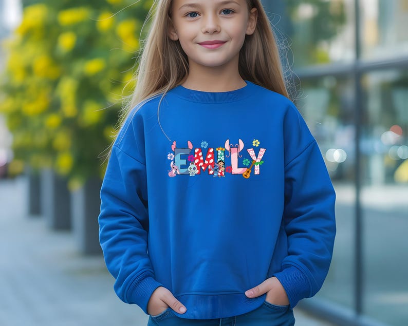 May include: A royal blue sweatshirt with the name "EMILY" in colorful, whimsical letters. The letters are decorated with various cartoon characters and floral accents. The sweatshirt is a long-sleeved, crew neck style.
