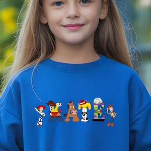 May include: A blue sweatshirt featuring the word "STATE" in a colorful, cartoon-style design. Each letter is styled after a character from a popular animated movie. The sweatshirt is worn by a person with long blonde hair.