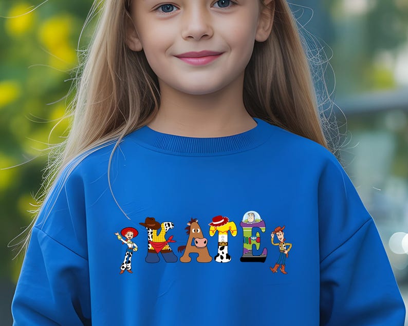 May include: A blue sweatshirt featuring the word "STATE" in a colorful, cartoon-style design. Each letter is styled after a character from a popular animated movie. The sweatshirt is worn by a person with long blonde hair.