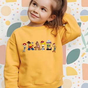 May include: A yellow sweatshirt featuring the name "KAILI" in colorful letters, each designed with characters from a popular animated movie. The sweatshirt has long sleeves and a crew neck. The background is a colorful pattern.
