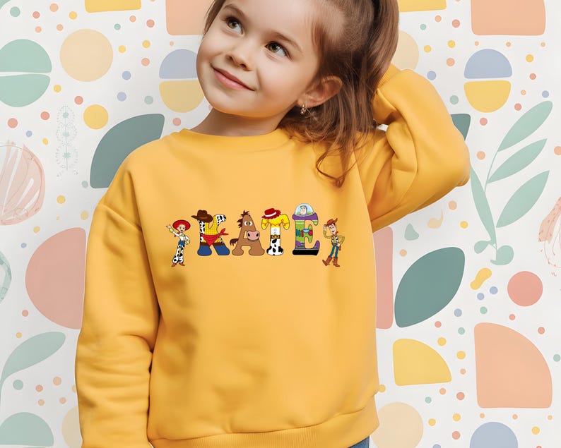 May include: A yellow sweatshirt featuring the name "KAILI" in colorful letters, each designed with characters from a popular animated movie. The sweatshirt has long sleeves and a crew neck. The background is a colorful pattern.
