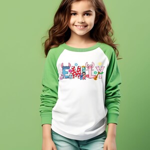 May include: A white and green long-sleeved shirt with the name "EMILY" printed across the front in colorful letters and cartoon characters. The shirt has a white body and green sleeves.