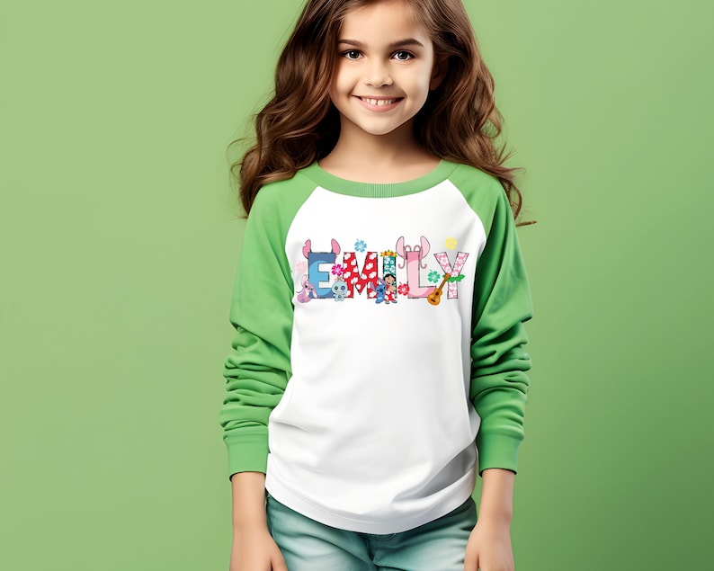 May include: A white and green long-sleeved shirt with the name "EMILY" printed across the front in colorful letters and cartoon characters. The shirt has a white body and green sleeves.