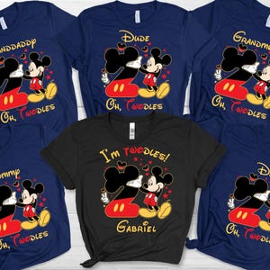 May include: A selection of navy blue and black t-shirts with a Mickey Mouse graphic. The design features the number 2 in red and yellow, with Mickey Mouse giving a peace sign. Text includes "Granddaddy", "Dude", "Grandmom", "Mommy", "Dad", and "I'm 2oodles!"