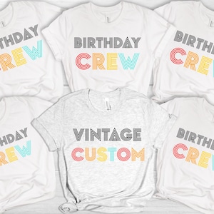 May include: A collection of white and grey t-shirts. The white shirts have "BIRTHDAY CREW" printed in black and rainbow hues. The grey shirt features "VINTAGE CUSTOM" in black and rainbow. These are casual, everyday wear t-shirts.
