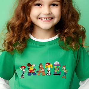 May include: A green t-shirt with white trim featuring the word "PEACE" spelled out with cartoon characters. The characters are from the Toy Story franchise. The shirt is worn by a young girl with long brown hair.