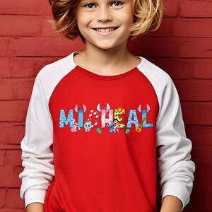May include: A red and white raglan-style shirt with the name "MICHAEL" printed across the chest in colorful, cartoon-style letters. The letters are decorated with various cartoon characters. The shirt is worn by a child.