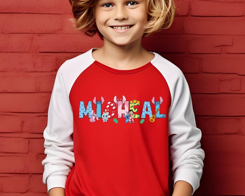 May include: A red and white raglan-style shirt with the name "MICHAEL" printed across the chest in colorful, cartoon-style letters. The letters are decorated with various cartoon characters. The shirt is worn by a child.