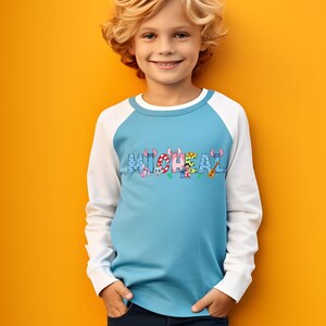 May include: A light blue and white raglan shirt with the name "MICHAEL" printed across the chest in colorful letters. The letters are decorated with cartoon characters and musical instruments. The shirt is worn by a child.