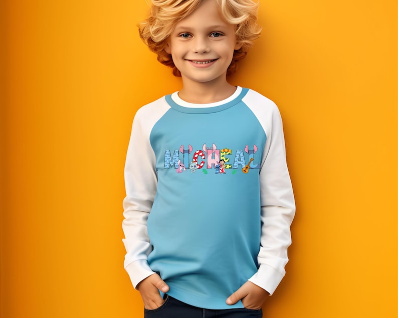May include: A light blue and white raglan shirt with the name "MICHAEL" printed across the chest in colorful letters. The letters are decorated with cartoon characters and musical instruments. The shirt is worn by a child.