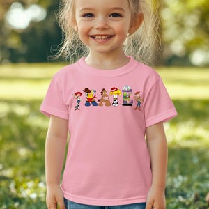 May include: A pink t-shirt featuring the word "CATTLE" in colorful cartoon letters, each designed with a different character from the animated movie. The shirt is worn by a young child, with a blurred green background.