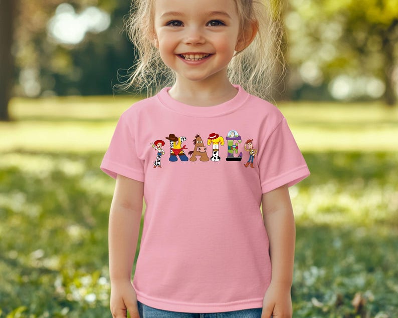 May include: A pink t-shirt featuring the word "CATTLE" in colorful cartoon letters, each designed with a different character from the animated movie. The shirt is worn by a young child, with a blurred green background.