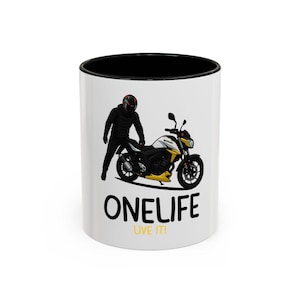 Peut inclure: Mug en céramique blanche avec intérieur noir. Il affiche les mots "ONELIFE LIVE IT!" en noir, ainsi qu'un dessin d'une moto et d'une personne en casque et tenue noire.