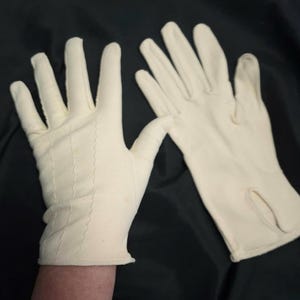 May include: A pair of off-white gloves. The gloves are made of a soft material and feature a stitched design on the back of the hand. The gloves have a classic design with a small slit on the side of one glove.