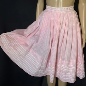 May include: A vintage pink and white gingham skirt. The skirt has a full, flared shape with pleats at the waist. The hemline is detailed with white horizontal stripes. The skirt is made of a lightweight fabric.