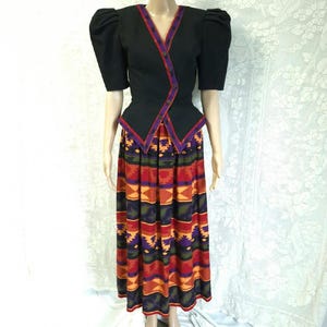 Peut inclure: Ensemble vintage deux pièces. La veste noire a des manches courtes bouffantes et un col en V bordé de rouge et de violet. La jupe est longue et fluide, avec un motif géométrique coloré dans les tons de rouge, orange, violet et vert.