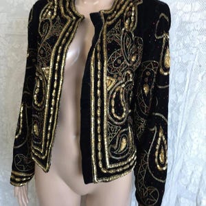 Peut inclure: Veste en velours noir ornée de sequins et de perles dorées. La veste à devant ouvert présente des motifs complexes et des motifs cachemire. Les manches longues et le col sont également ornés de sequins et de perles dorées.