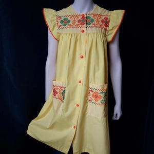 Könnte beinhalten: Ein gelbes Kleid mit floraler Kreuzstichstickerei auf dem Oberteil und den Taschen. Das Kleid hat kurze Ärmel mit orangefarbenem Besatz und orangefarbenen Knöpfen vorne. Das Kleid wird auf einer Schaufensterpuppe präsentiert.