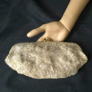 May include: A cream-colored clutch bag covered in intricate beadwork and sequins, featuring a floral-inspired pattern across the surface. The bag has a gold-toned metal kiss-lock clasp closure at the top center, providing a secure opening mechanism.