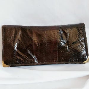 Puede incluir: Un bolso de mano marrón oscuro con un patrón texturizado similar al de un reptil. El bolso rectangular tiene un diseño plegable y detalles dorados en las esquinas. El bolso de mano está sobre una superficie blanca.