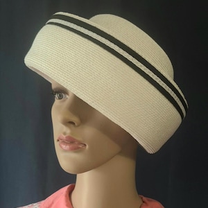 May include: A cream-coloured straw hat with a flat top and a slightly angled brim. The hat features two black bands around the crown. The hat is displayed on a mannequin head, showcasing its shape and design. The hat is a vintage style.