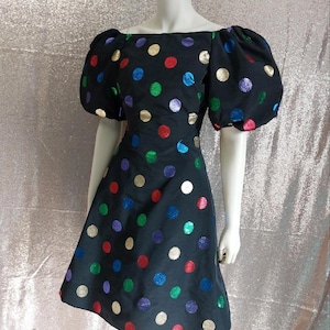 May include: A black dress with large, colorful polka dots in gold, red, green, blue, and purple. The dress features an off-the-shoulder neckline and puffed sleeves. The dress is displayed on a mannequin.