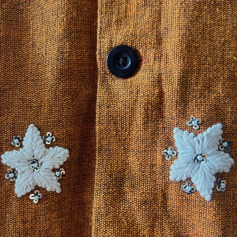 May include: Close-up of a textured orange fabric with a black button and two white embroidered flower designs. The flowers have silver sequins and are centered with a small silver bead.