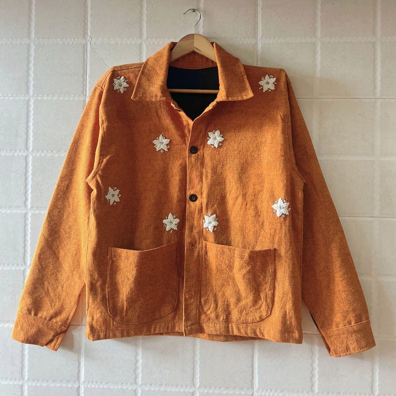 May include: An orange jacket with long sleeves, two front pockets, and black buttons. The jacket is decorated with white, star-shaped embellishments. The jacket is hanging on a wooden hanger against a white textured background.