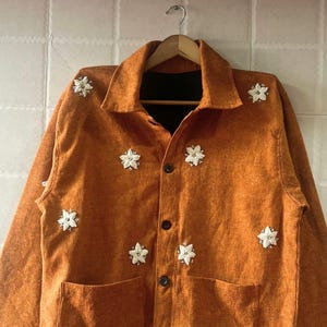 May include: An orange jacket with a collared neckline and two front pockets. The jacket features decorative white floral embellishments and black buttons down the front. The jacket is hanging on a wooden hanger.