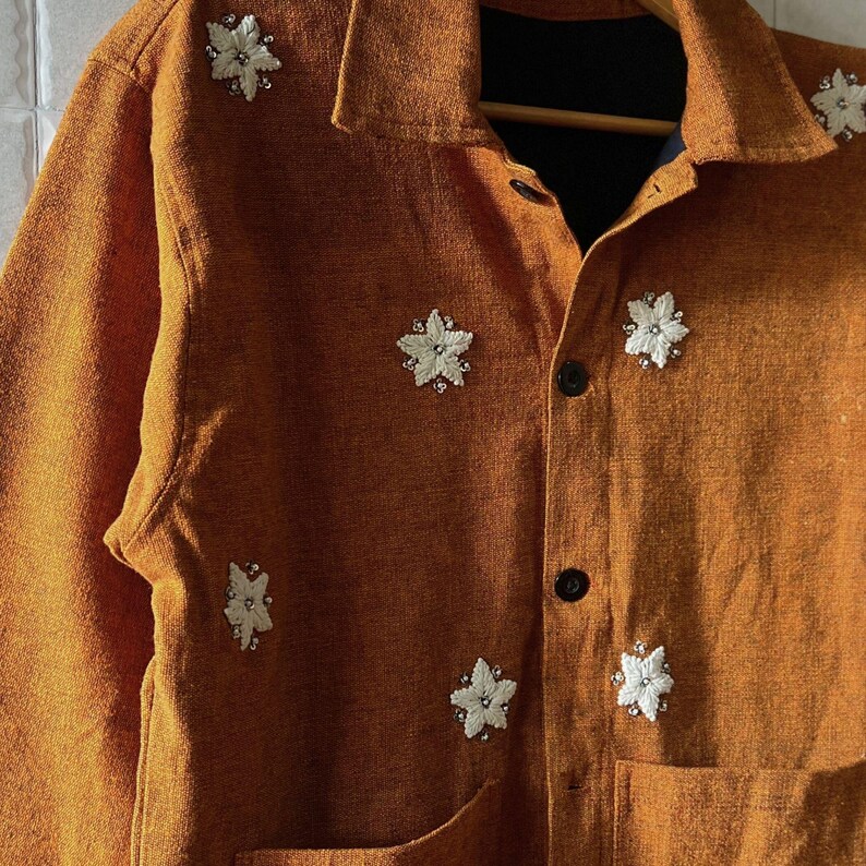 May include: An orange-brown jacket with a collared neckline and two front pockets. The jacket is adorned with multiple white floral embellishments with crystal-like centers. The jacket has black buttons and is hanging on a wooden hanger.