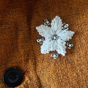 May include: Close-up of a textured orange fabric with a large, white, embroidered flower and small, decorative embellishments. A black button is visible on the lower left.