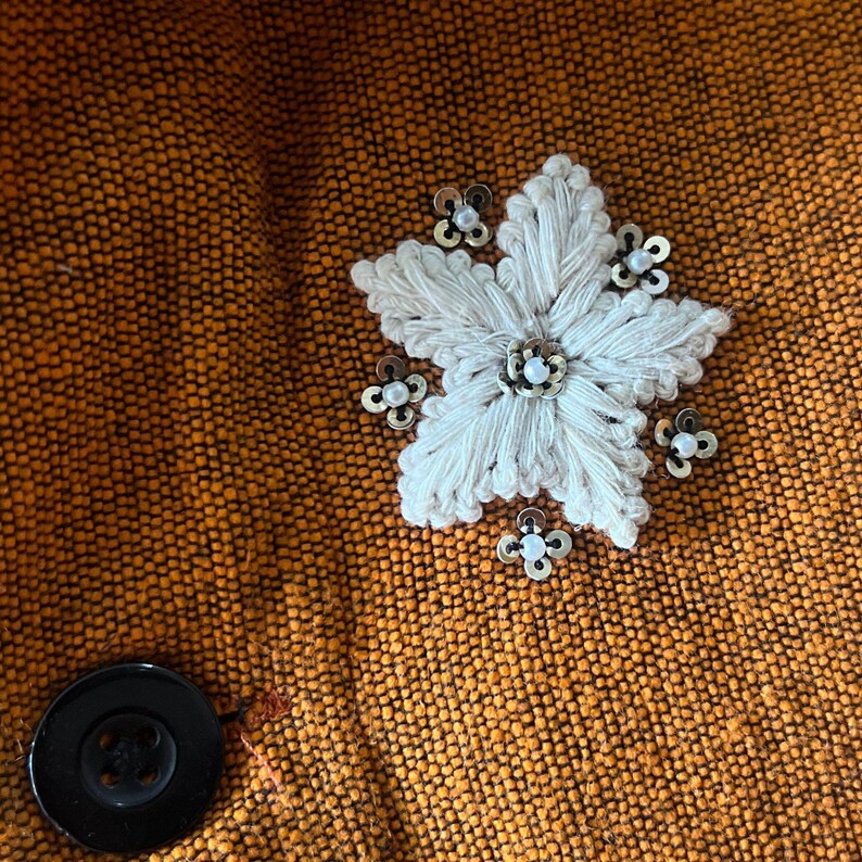 May include: Close-up of a textured orange fabric with a large, white, embroidered flower and small, decorative embellishments. A black button is visible on the lower left.