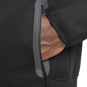 Puede incluir: Primer plano de una sudadera negra con un bolsillo con cremallera gris. La cremallera est&aacute; parcialmente abierta, revelando una mano en el interior. La sudadera tiene un aspecto texturizado y un cord&oacute;n en la parte inferior de la cremallera.