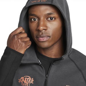 Puede incluir: Una sudadera con capucha gris oscuro con el texto "LIFE OF SYN" en la capucha. La sudadera tiene una cremallera y un dise&ntilde;o gr&aacute;fico en naranja y rosa en el pecho. El logotipo de Nike es visible en la parte inferior.