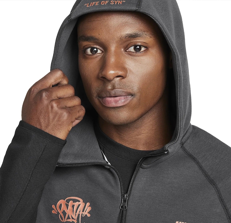 Puede incluir: Una sudadera con capucha gris oscuro con el texto "LIFE OF SYN" en la capucha. La sudadera tiene una cremallera y un dise&ntilde;o gr&aacute;fico en naranja y rosa en el pecho. El logotipo de Nike es visible en la parte inferior.