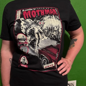 May include: Black t-shirt featuring a comic book-style graphic of the Mothman. The design includes the text "The Diabolical Mothman" and a red car. Additional text includes "It seemed to be waiting on us."