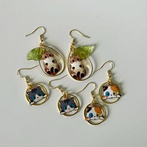 May include: Five gold-toned dangle earrings featuring enamel cat designs. Two earrings have teardrop frames with green leaves and brown cats. Three earrings have circular frames with gray or calico cats peeking over the bottom edge.