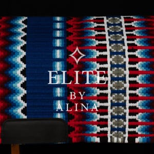 May include: A blue, red, and white woven saddle pad with a geometric pattern. The pad features a zig-zag design and vertical stripes. The text "ELITE BY ALINA" is displayed in white. The pad is designed for equestrian use.