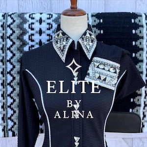 May include: A black long-sleeve show shirt with white piping, featuring a collar and cuffs heavily embellished with clear crystals and silver accents. The text ELITE BY ALINA is centered on the front. A black, white, and gray patterned saddle pad is visible behind the shirt.