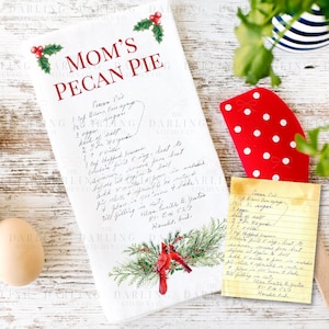 May include: A white kitchen towel with the text "MOM'S PECAN PIE" in red, along with a handwritten recipe. The towel features holly and cardinal illustrations. Eggs and a red spatula with white polka dots are also visible.